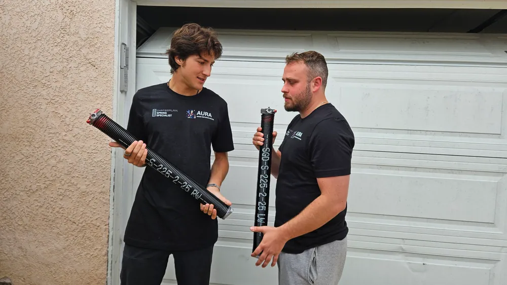 Two Aura technicians comparing torsion springs outside a garage