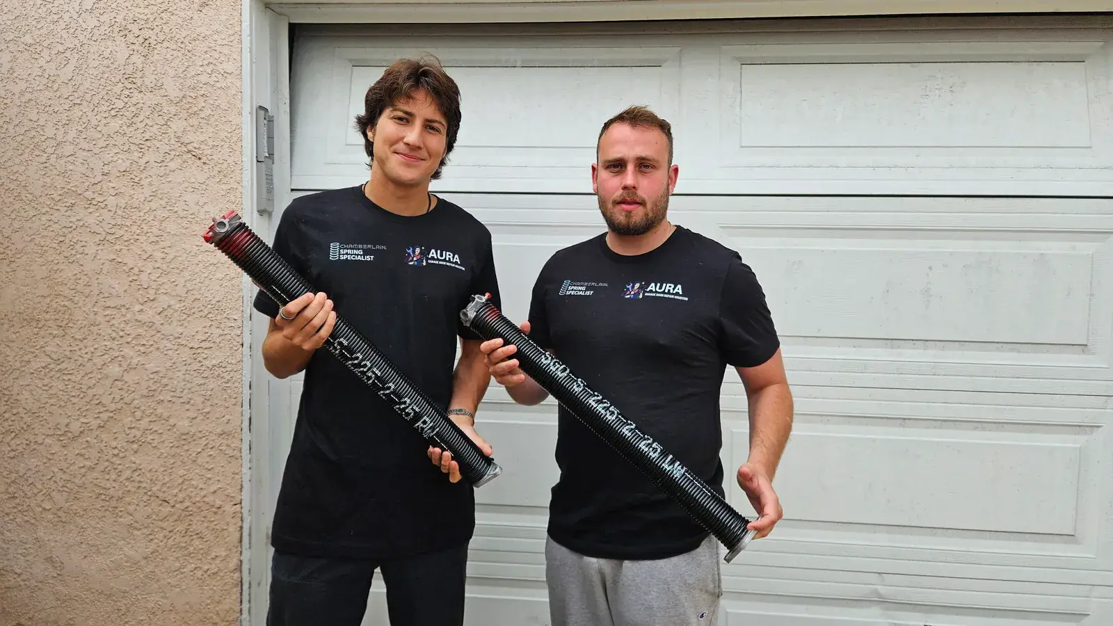 Two Aura technicians holding torsion springs in front of a white garage door